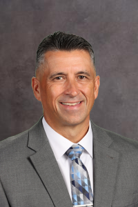 Professional head-and-shoulders portrait of a smiling man in a gray suit and blue patterned tie, neutral backdrop.