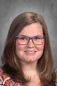 Portrait of a smiling woman with shoulder-length brown hair and purple glasses against a gray backdrop, in a professional headshot style.