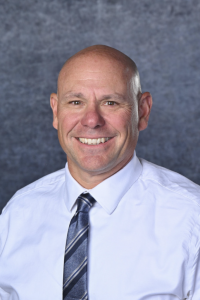 Smiling bald man in a white dress shirt and striped tie against a blue backdrop (professional headshot).
